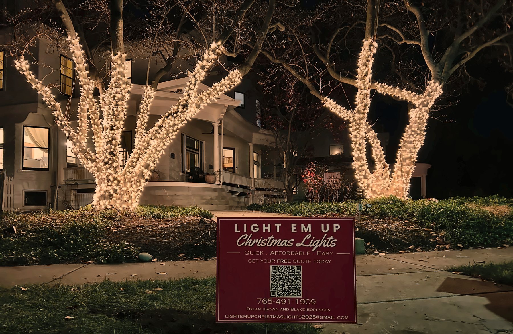 Two large trees wrapped trunk to branch in warm white lights in front of a historic home, with the Light 'Em Up yard sign